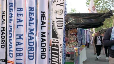 Real-Madrid-football-team-scarves-are-seen-selling-at-a-street-stall-as-fans-walk-past-the-frame-before-attending-the-Champions-League-football-match-against-Manchester-City-football-team