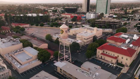 Aerial-View-of-WB-Water-Tower-at-Warner-Brothers-Studio-Lot-in-Burbank-California,-Drone-Shot-Flying-Over-Backlot-and-Soundstages-in-Daytime