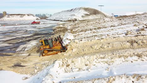 Yellow-bulldozer-removing-snow-and-soil-on-slope,-construction-in-Montreal,-Quebec,-Canada