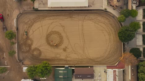 Drone-Sobre-Caballos-Con-Entrenadores-En-Corral-Ecuestre,-El-Centro-Ecuestre-Durante-El-Día-Visto-Desde-El-Aire