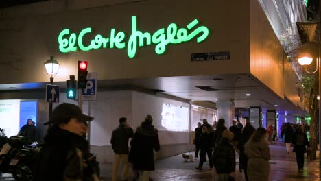 People-walk-across-the-street-in-front-of-Spain's-largest-department-store,-El-Corte-Inglés,-at-nighttime