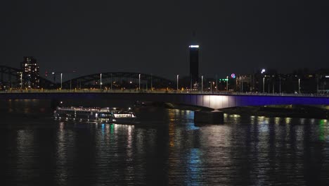 Embark-on-a-mesmerizing-journey-along-the-Rhine-River-by-night,-as-a-cruise-ship-glides-gracefully-past-Cologne's-illuminated-bridges,-creating-a-captivating-spectacle