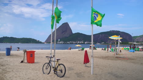 A-vista-of-Copacabana-Beach-in-Rio-de-Janeiro,-Brazil,-reveals-locals-relaxing-amidst-two-Brazilian-flags-and-a-bicycle,-against-the-backdrop-of-a-mountain-range-and-expansive-blue-skies