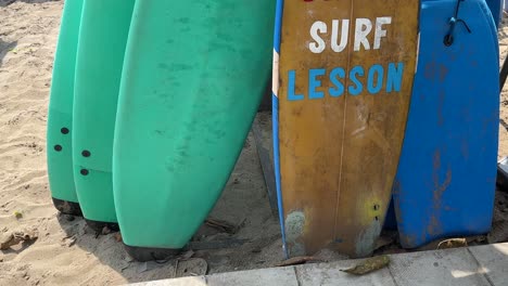 Surf-Lesson-painted-on-a-surfboard-amongst-a-group-of-surfboards-resting-in-the-sand-of-Legian-Beach,-Bali,-Indonesia