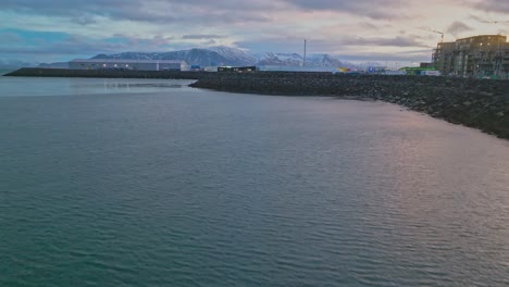 Drone-video-flying-along-Iceland's-Reykjavik-City-Waterfront-rising-to-reveal-a-moody-sunrise-over-snow-capped-mountain-range