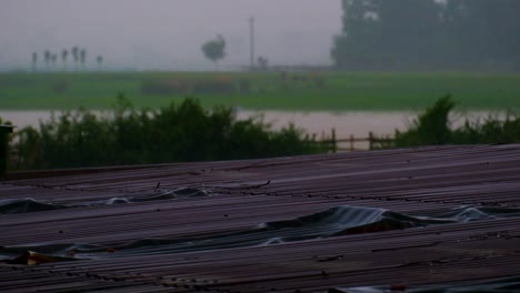 Strömender-Regen-Auf-Dem-Rustikalen-Dach-Eines-Ländlichen-Blechschuppens-Am-Monsunabend-Am-Flussufer-Mit-Ruhiger-Dorfkulisse