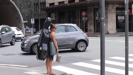 Una-Mujer-Músico-Con-Un-Vialonchell-En-La-Espalda-Cruza-La-Calle-En-Un-Tráfico-De-Autos-En-Génova,-Italia