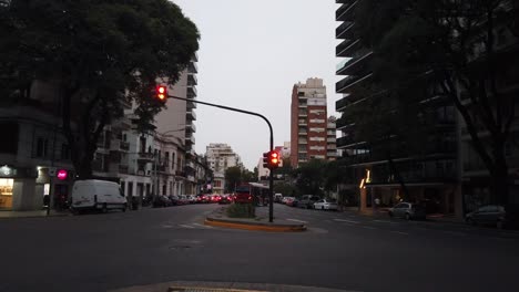 Skyline-Bei-Sonnenuntergang,-Busse-Mit-öffentlichen-Verkehrsmitteln-Fahren-An-Der-Ampel-Der-Stadtarchitektur-Von-Buenos-Aires-Vorbei,-Menschen-Mit-Einrädern-Laufen-An-Der-Belebten-Straßenkreuzung-Im-Viertel-Alberdi-Flores-Vorbei