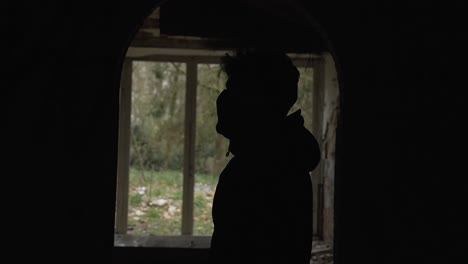 Young-man-silhouette-standing-with-black-hoodie-inside-corridor-and-in-front-of-big-window-in-abandoned-mansion