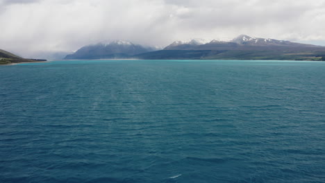 Fliegen-Sie-über-Das-Strahlend-Blaue-Wasser-Des-Lake-Pukaki,-Während-Tiefe-Wolken-Die-Umliegenden-Schneebedeckten-Malerischen-Berge-Verhüllen