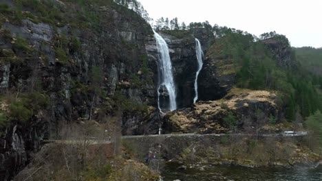 Tourists-Standing-next-to-see-and-looking-towards-Hesjedalsfossen-waterfall,-reverse-aerial