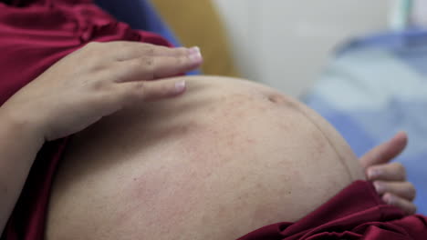 Schwangere-Frau-Trägt-Einen-Roten-Seidenpyjama-Und-Liegt-Auf-Dem-Bett,-Während-Sie-Mit-Beiden-Händen-Sanft-Ihren-Geschwollenen-Bauch-Streichelt
