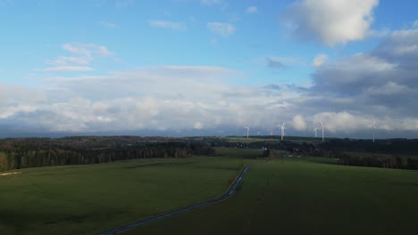 Aerialview-of-scenic-view-of-a-green-field-with-a-winding-path,-bordered-by-trees-and-wind-turbines-under-a-partly-cloudy-sky