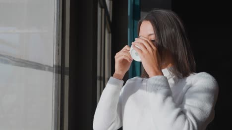 Young-female-stand-near-apartment-window-and-enjoy-morning-coffee-in-sunlight