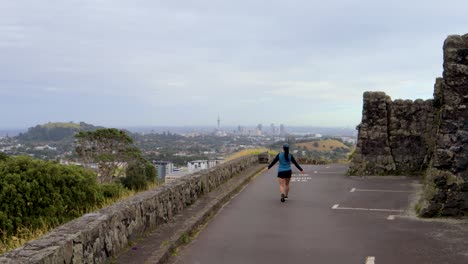 Frau-Joggt-Und-Hebt-Ihre-Arme-In-Felsiger-Mittelalterlicher-Architektur,-Nebliger-Landschaft-Oben-Auf-One-Tree-Hill,-Auckland-Skyline-Hintergrund