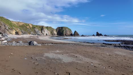 Menschen,-Die-Mit-Hunden-Am-Strand-Spazieren-Gehen,-Mit-Blauem-Himmel-Und-Weißen-Wellen,-Die-Im-Frühling-An-Den-Strand-In-Waterford,-Irland,-Plätschern