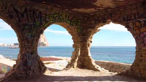 Peñon-D&#39;ifach---También-Conocido-Como-Roca-De-Calpe:-Espectacular-Vista-De-Drones-FPV-De-La-Icónica-Montaña-Y-Costa-De-Calpe-España-A-Través-De-Ventanas-Circulares-Pintadas-En-Un-Edificio-De-Graffiti-Abandonado