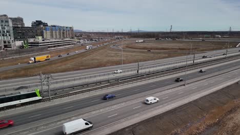 Aerial-of-Réseau-express-métropolitain-REM-automated-light-rail-system-in-Brossard,-near-Montreal-city,-with-car-traffic-on-the-main-highway-,-modern-infrastructure-low-gas-emission-pollution