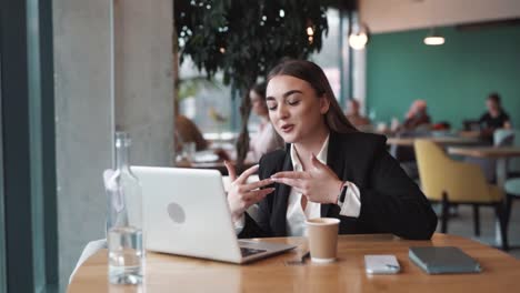cheerful,-beautiful-woman-sits-in-a-stylish-cafe-in-business-attire-with-a-laptop,-joyfully-conducting-training-and-conference-calls-via-video-link-with-a-smile