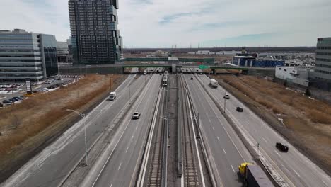 aerial-front-view-of-The-Réseau-express-métropolitain-REM-automated-light-rail-system-in-Brossard-near-Montreal-city,-driving-along-the-main-highway-with-car-traffic
