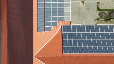 Aerial-view-of-a-building-with-a-terracotta-tiled-roof-partially-covered-with-rows-of-blue-solar-panels,-adjacent-to-a-paved-area-with-shadows