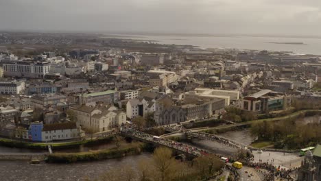 Eine-Drohne-Steigt-über-Die-Zuschauer-Der-Parade-Auf,-Die-Die-Seltsame-Salmon-Bridge-über-Den-Fluss-Corrib-In-Galway,-Irland-überqueren.