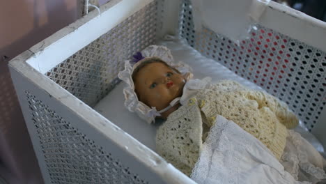 Antique-creepy-doll-in-a-vintage-wire-crib,-adorned-with-lace-and-crochet-blanket,-capturing-a-bygone-era