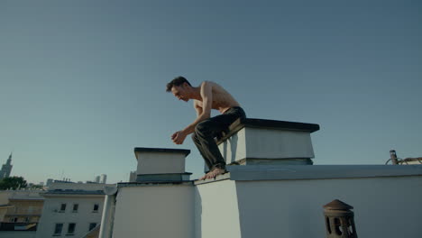 Young-bare-chested-man-sits-down-on-roof-of-city-building-in-sunlight,-handheld
