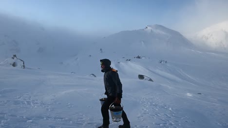 Man-carrying-buckets-of-water-uphill-through-blizzard-towards-mountain-cabin
