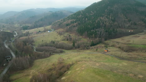 An-aerial-view-shows-a-winding-road-cutting-through-a-verdant-valley-with-a-solitary-orange-roofed-house-nestled-amid-rolling-hills-and-dense-woodlands