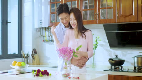 Happy-Young-Couple-Hug-and-Making-Bouquet-Flowers-On-A-Wooden-Table-In-Kitchen-Together-for-anniversary,-valentines-day-or-birthday