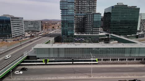 aerial-established-of-The-Réseau-express-métropolitain-REM-automated-light-rail-system-in-Brossard,-near-Montreal-city,-with-car-traffic-on-the-main-highway-and-skyline-modern-buildings-cityscape