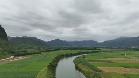 Aerial-Drone-Shot-of-River-Passing-Through-Crop-Fields-With-Rain-Clouds-In-Sky