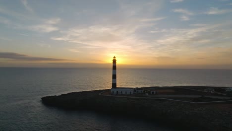 Golden-sun-shine-over-Artux-Lighthouse-Menorca-landmark-beach-shore-drone-aerial-coastal-islet,-calm-ocean-water-at-Balearic-islands