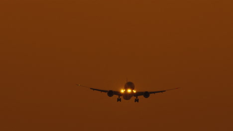 Frontal-view---an-airplane-with-headlights-on-comes-in-to-land-at-an-airport-and-lands-on-a-runway-airstrip-at-sunset-with-orange-coloured-sky