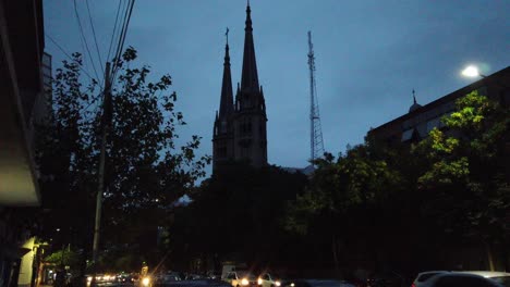 City-Skyline-at-night-Basilica-Our-Lady-of-Buenos-Aires-Tall-Tower-over-blue-sky-cars-driving-by-nighttime-fast-lane,-latin-metropolitan-town-religious-landmark