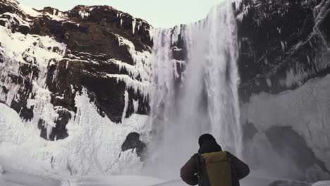 tourist-walking-in-slow-motion-close-to-Skogafoss-waterfall-in-winter-season,-with-water-and-ice-scenic-landscape