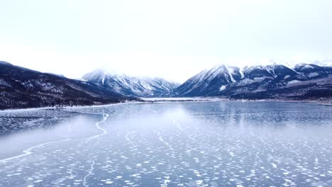 Hoch-über-Einem-Zugefrorenen-See-Weg-Von-Schneebedeckten-Bergen-Im-Winter-Fliegen,-Luft