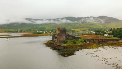 Wide-Aerial-Pan-of-Eilean-Donan,-a-Famous-Castle-on-Loch-Duich,-Revealing-Cloudy-Scottish-Highlands-in-Autumn,-Scotland,-United-Kingdom