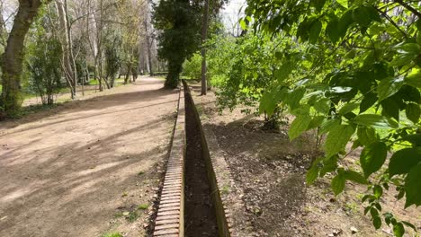 Vemos-Las-Imágenes-De-Un-Canal-De-Riego-De-Ladrillo-Sobre-Un-Camino-Arenoso,-Por-Un-Lado-árboles-Dando-Sus-Primeros-Brotes-Y-Por-El-Otro-Con-Hojas-Verdes-En-El-Jardín-Del-Príncipe-Aranjuez,-España