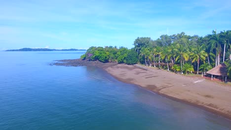 An-empty-island-white-sand-beach-with-house-and-small-thatched-sun-hut-lined-with-palm-trees-and-calm-ocean-water