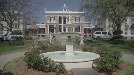 A-wide-shot-of-a-grand-historical-building-in-Nashville,-with-a-fountain-in-the-foreground-surrounded-by-well-maintained-gardens-and-pathways,-under-a-clear-sky