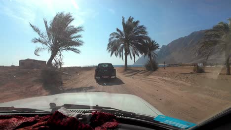 A-white-vehicle-in-Egypt-with-a-red-and-black-scarf-draped-over-the-dashboard-is-driving-on-a-dirt-road-toward-the-ocean,-with-a-black-pickup-truck-ahead,-several-palm-trees-lining-the-path