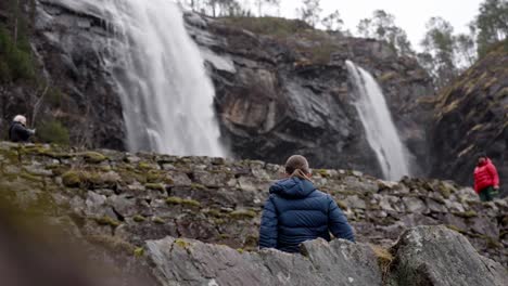 Teen-girl-watching-Norway-waterfall,-Cinematic-reveal-from-behind-rock