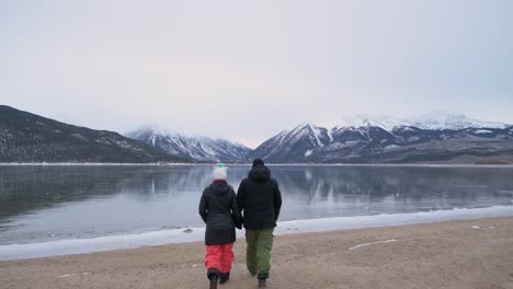 Couple-holding-hands-walking-to-the-edge-of-a-frozen-lake-with-snow-covered-mountains-in-the-background,-static