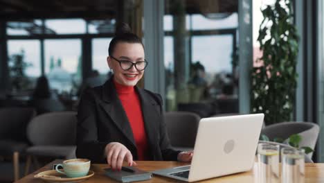 beautiful-young-woman-in-glasses-and-braces,-a-freelancer,-sits-in-a-stylish-restaurant-with-a-laptop,-dressed-in-business-attire-and-glasses,-smiling-happily-at-the-news-of-winning-and-victory