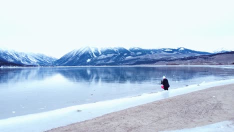 Photographer-on-the-edge-of-a-frozen-lake-with-snow-covered-mountains-in-the-background,-aerial