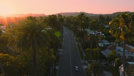 Aerial-Establishing-Shot-of-Sunlit-Beverly-Hills-Neighborhood-at-Sunset,-Golden-Sun-Above-and-Upscale-Homes-Below,-Cars-Driving-Along-Iconic-Street-with-Mountains-on-Horizon