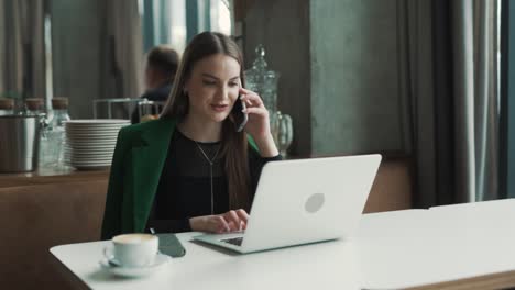 beautiful-young-woman,-a-freelancer,-sits-in-a-stylish-restaurant-dressed-in-business-attire,-talking-on-the-phone-and-working-on-her-laptop