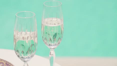 Zwei-Tassen-Weißwein-Und-Champagner-Auf-Einem-Tisch-Mit-Klarem-Wasser-Im-Swimmingpool-Im-Hintergrund